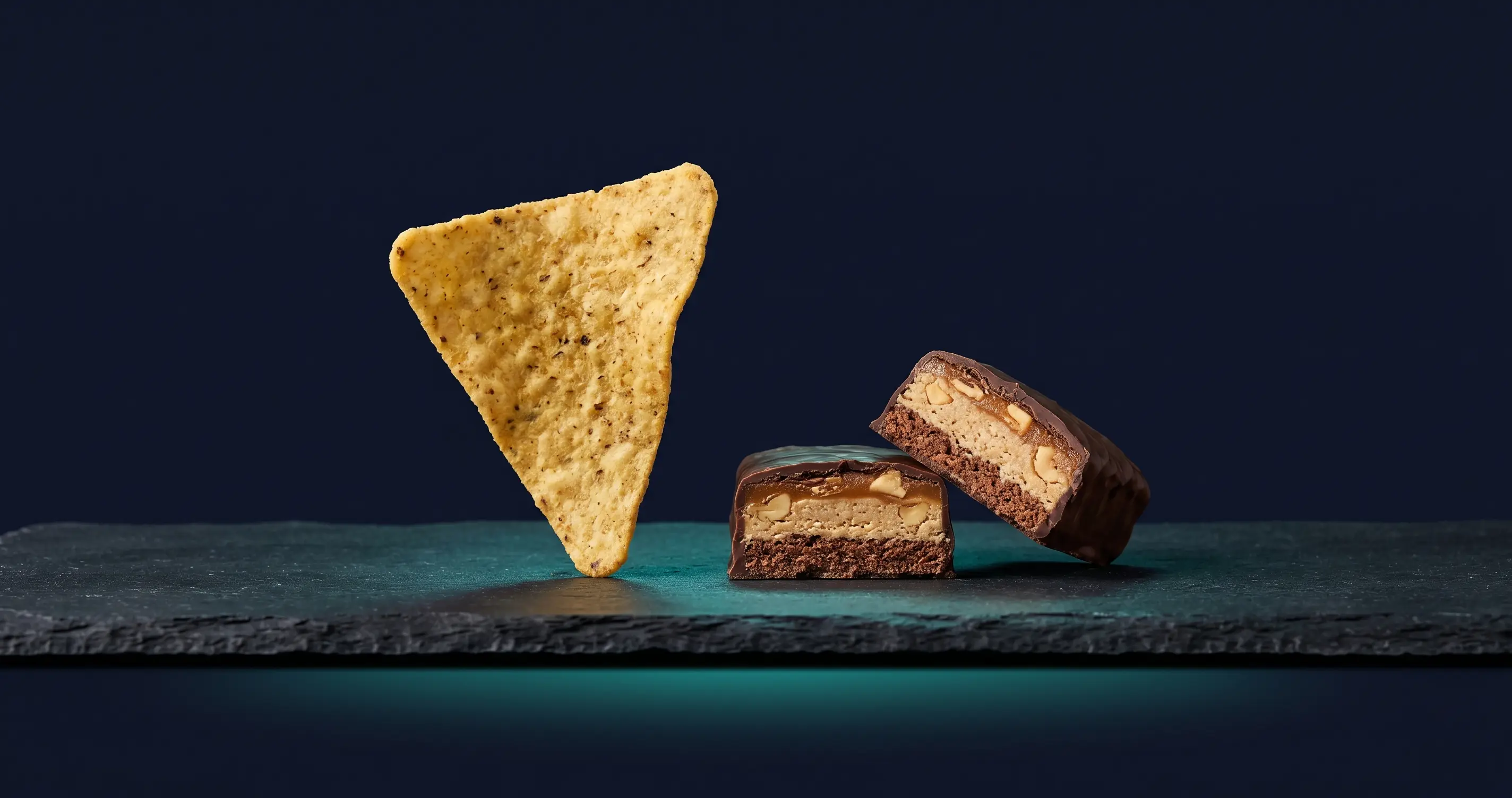 A tortilla-style protein chip balancing on its edge next to a protein bar cut in half, on a dark studio surface.